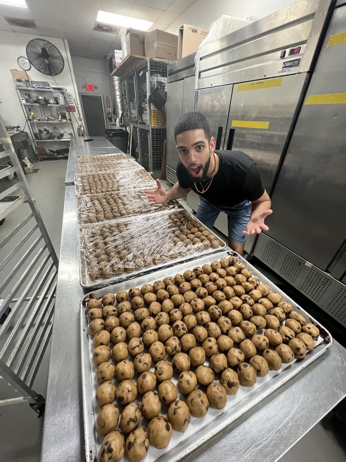 Mike, owner of mmmbaked, grinning in his commercial kitchen with both arms out over full trays of raw chocolate-chip cookie dough and wrapped finished cookies